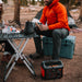 Image of Jackery Explorer 500 portable power station used during outdoor camping, positioned on the ground powering an electric kettle on a folding table with people seated nearby in a forest campsite setting.