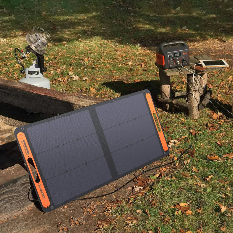 Image of Jackery solar panel connected to Jackery Explorer portable power station outdoors, angled front view of foldable solar panel with power station and devices set on wooden stands in a grassy campsite setting.
