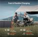 Image of Jackery Explorer 600 portable power station being charged outdoors with solar panel while two people work at a camping table under a canopy, dark gray unit with orange accents, lifestyle scene in a grassy field with mountains in the background and charging information text overlay.