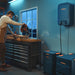 Image of Jackery home battery system with wall-mounted unit and expansion batteries powering workshop tools, black and orange units connected and in use, angled side view in indoor garage setting.