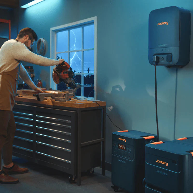 Image of Jackery home battery system with wall-mounted unit and expansion batteries powering workshop tools, black and orange units connected and in use, angled side view in indoor garage setting.