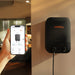 Image of Jackery Smart Transfer Switch wall-mounted in a home interior with black casing and orange logo, shown alongside a smartphone displaying energy monitoring app, angled side view in indoor setting.