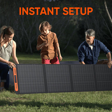 Image of a Jackery portable foldable solar panel being set up outdoors by two adults and a child on grass, black photovoltaic panels with orange accents, front view in a natural outdoor setting.