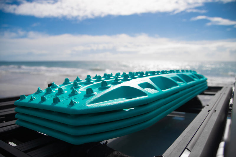 Image of stacked teal MAXTRAX MKII recovery traction boards mounted on a vehicle roof rack, angled close-up view with ocean beach background.