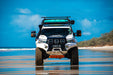 Image of off-road 4WD vehicle driving on beach with teal MAXTRAX MKII recovery boards mounted on roof rack, front view with ocean and sky background.