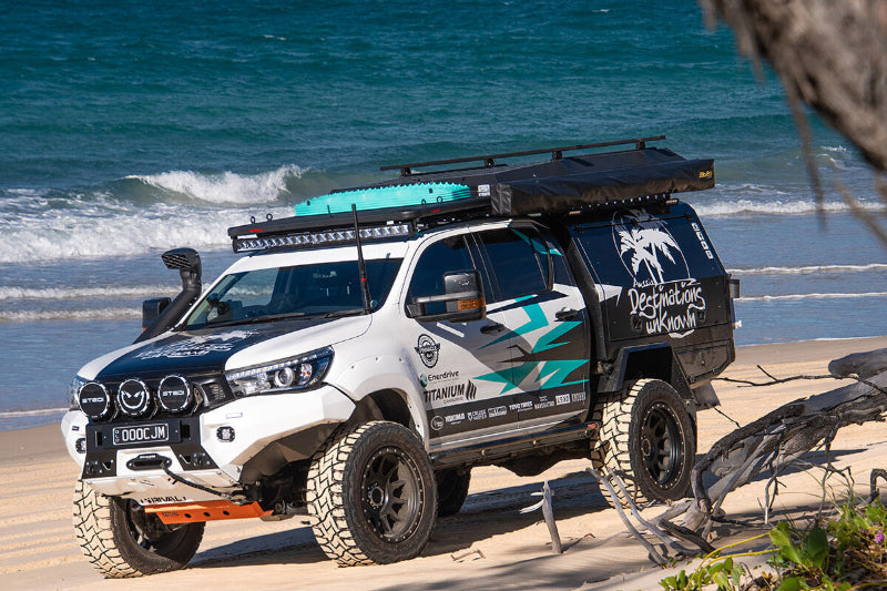 Image of off-road 4WD vehicle parked on sandy beach with teal MAXTRAX MKII recovery boards mounted on roof rack, angled front and side view with ocean background.