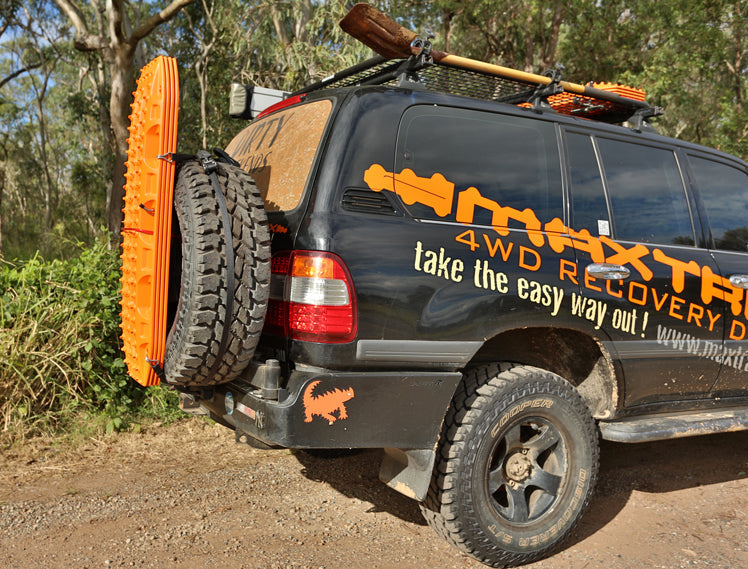 Black 4x4 vehicle with "MAXTRAX 4WD RECOVERY" decal, featuring orange MAXTRAX recovery boards mounted on the rear spare tire and a shovel on the roof rack, parked outdoors.