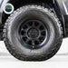 Image of a black off-road vehicle tire with a black rim, featuring an Up Down Air green valve stem deflator installed, on a white background with the "Overland Vehicle Systems" logo.