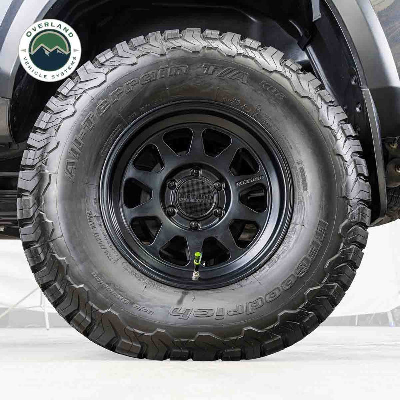 Image of a black off-road vehicle tire with a black rim, featuring an Up Down Air green valve stem deflator installed, on a white background with the "Overland Vehicle Systems" logo.
