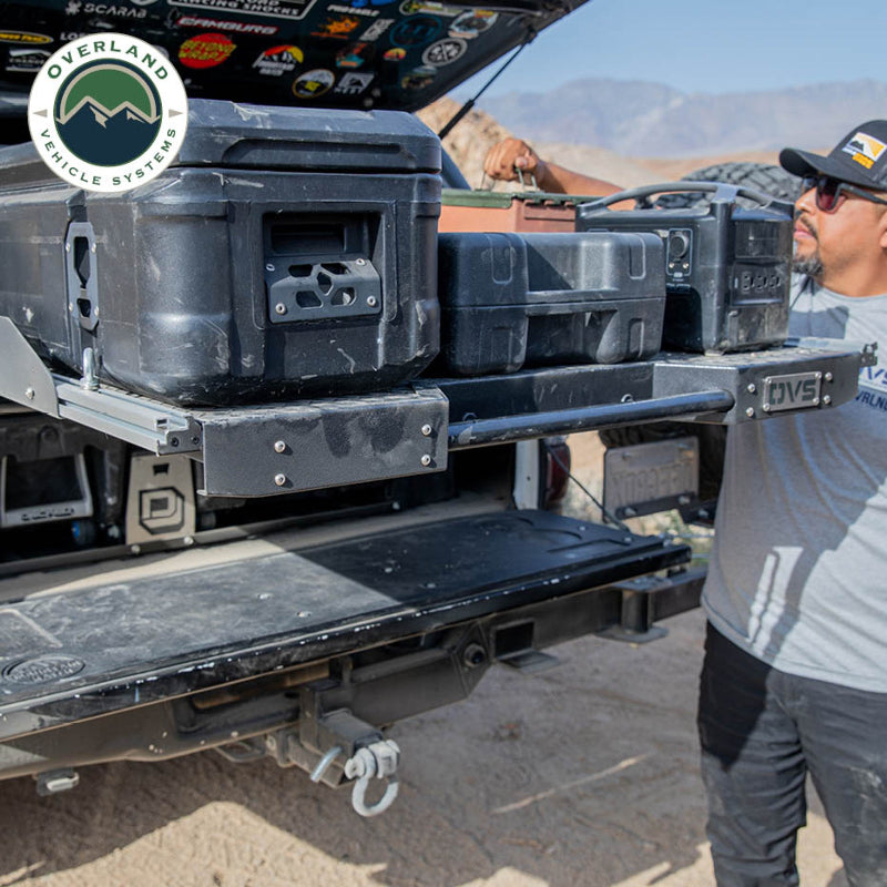 Image of Overland Vehicle Systems truck bed extension with storage cases, showing a person lifting a storage container, side view with a rugged outdoor background and dirt on the truck bed