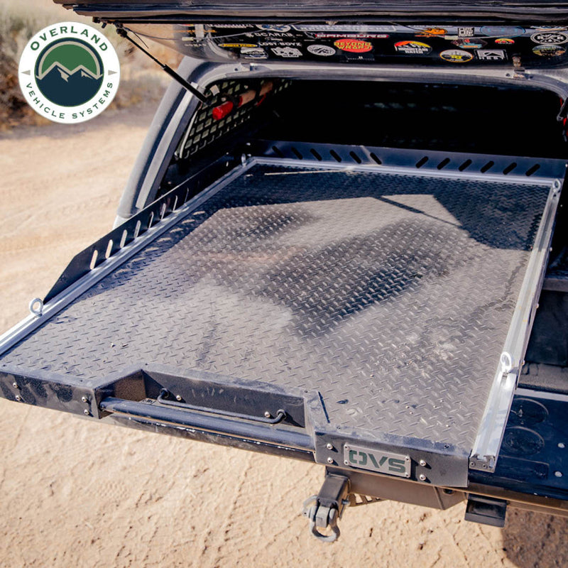 Image of Overland Vehicle Systems truck bed extension with diamond plate surface, partially extended, showing rugged outdoor terrain and the truck's hitch, side view with a focus on the sturdy extension system