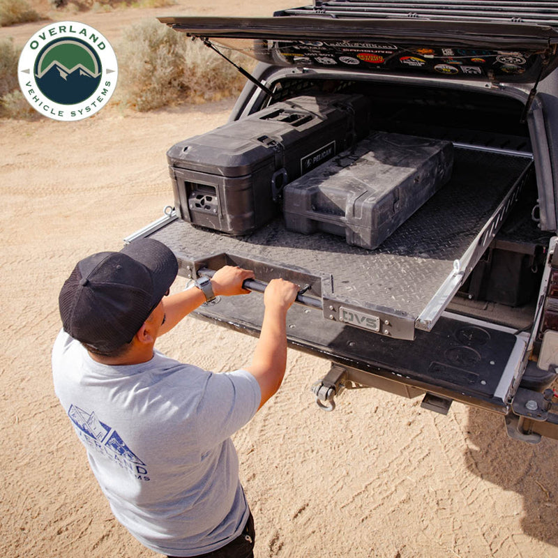 Image of Overland Vehicle Systems truck bed extension with diamond plate surface, showing a person pulling a Pelican storage case from the bed, side view with rugged terrain in the background