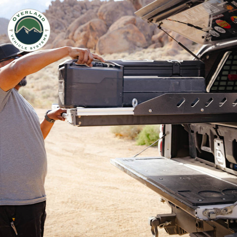 Image of Overland Vehicle Systems truck bed extension with a person lifting a storage container, showing the extension system with diamond plate surface and a rugged outdoor background