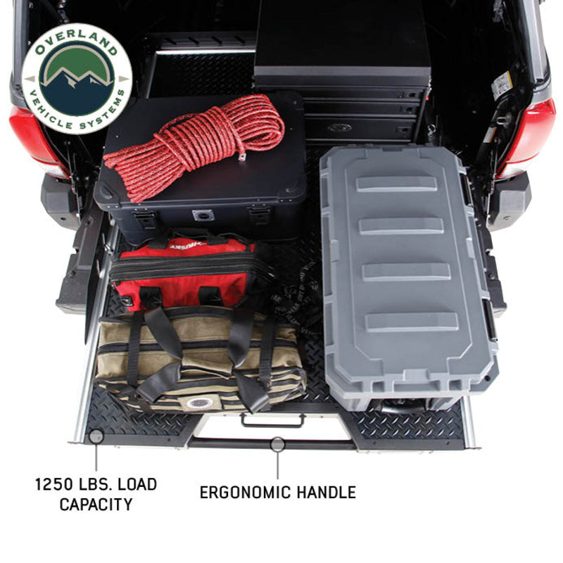 Image of Overland Vehicle Systems truck bed extension with diamond plate surface, showing storage boxes, rope, and bags, with text labels highlighting 1250 lbs load capacity and ergonomic handle, top-down view
