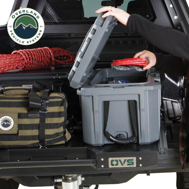 Image of Overland Vehicle Systems truck bed extension with storage box, showing a person removing a rope from the container, with a striped duffel bag placed nearby, side view on a diamond plate surface