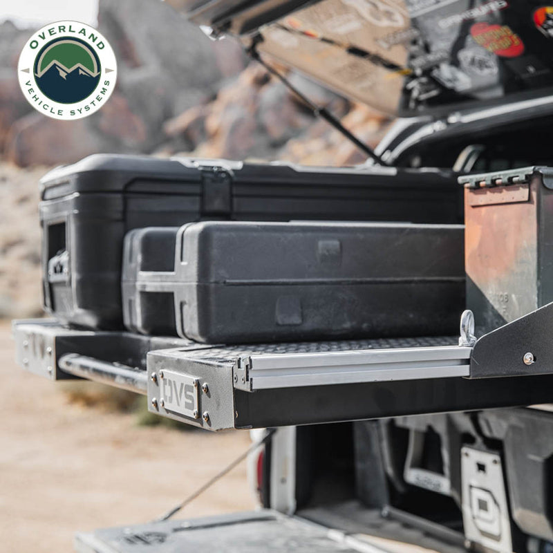 Image of Overland Vehicle Systems truck bed extension with storage cases, featuring a pull-out handle and diamond plate surface, side view with a rugged outdoor background