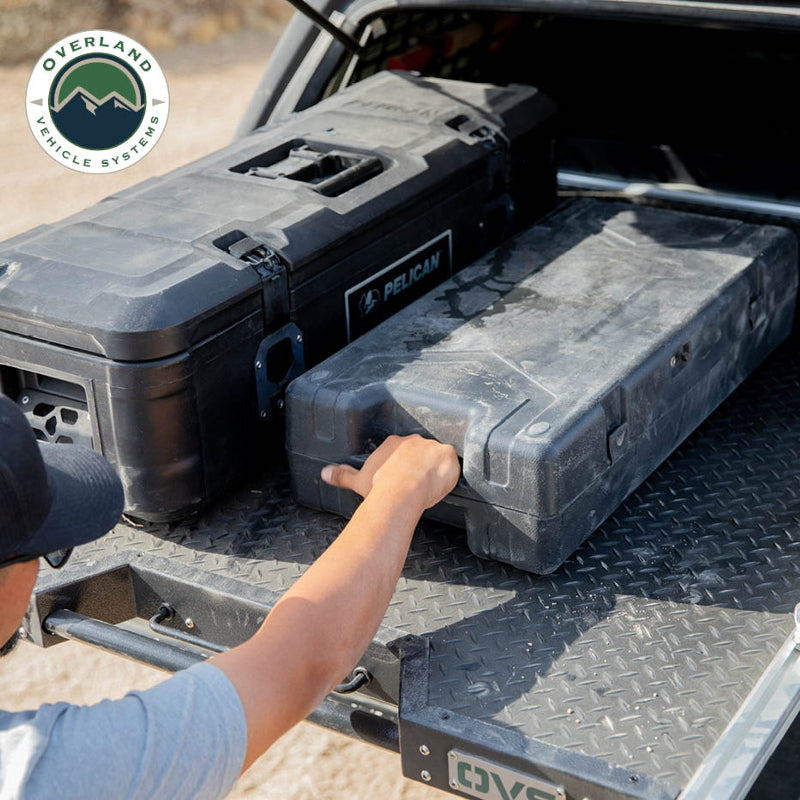 Image of Overland Vehicle Systems truck bed extension with diamond plate surface, showing a person pulling a Pelican storage case, side view with rugged outdoor background