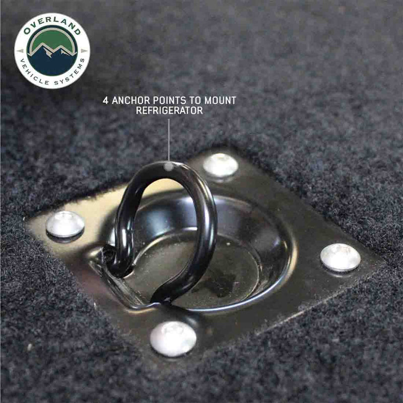 Image of a close-up on a black D-ring anchor point mounted on a dark gray marine-grade carpet, part of an Overland Vehicle Systems (OVS) Cargo Box, with text indicating "4 ANCHOR POINTS TO MOUNT REFRIGERATOR." The OVS logo is in the top left, all on a white background