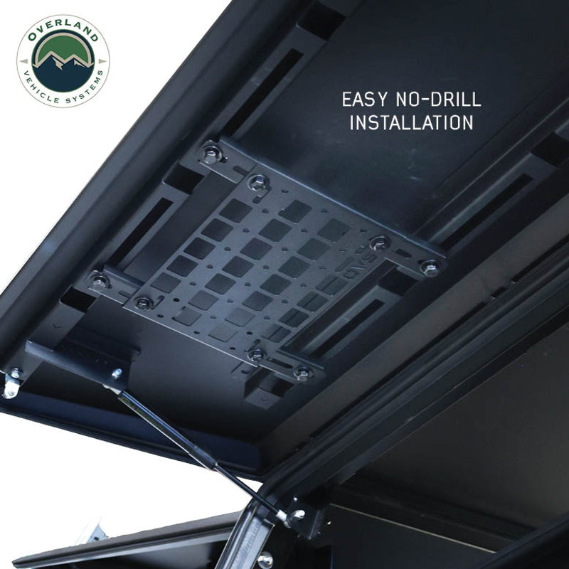 Image of a MOLLE panel installed on the underside of a truck cap or vehicle roof, demonstrating the no-drill installation with the included hardware. The panel is shown from a low angle, highlighting how it attaches to the existing structure