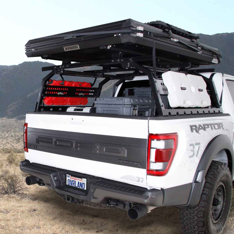 Image of Overland Vehicle Systems roof rack mounted on a Ford Raptor truck, featuring a rooftop tent, fuel canisters, and storage boxes in the truck bed, with a scenic background