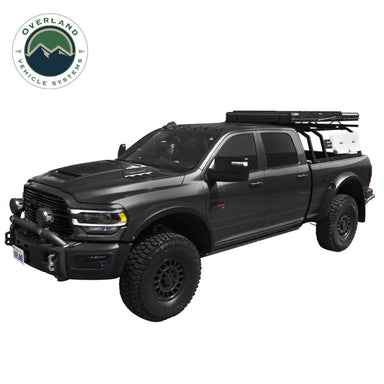 Image of a black full-size pickup truck with oversized tires, featuring a roof rack with gear mounted on top. The truck has an aggressive off-road appearance, with a front bull bar, fog lights, and a rugged design, viewed from the side on a white background