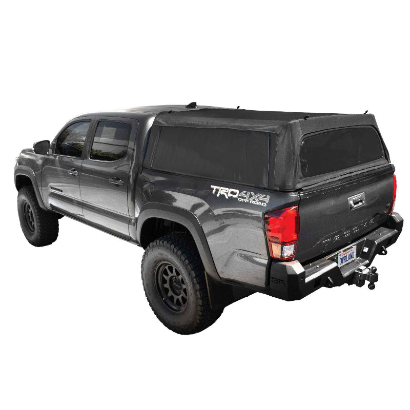 Image of an Overlanding Vehicle Systems Explorer Soft Shell bed topper on a dark gray Toyota Tacoma pickup truck, angled rear and side view on a white background. The soft shell topper is made of black diamond canvas with clear vinyl windows