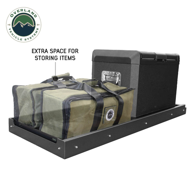 Image of an OVS black steel refrigerator slide holding a portable refrigerator and two green storage bags, with text "EXTRA SPACE FOR STORING ITEMS" pointing to the bags, all on a white background. This setup demonstrates how the slide can efficiently organize and secure both refrigeration and additional gear for overlanding adventures