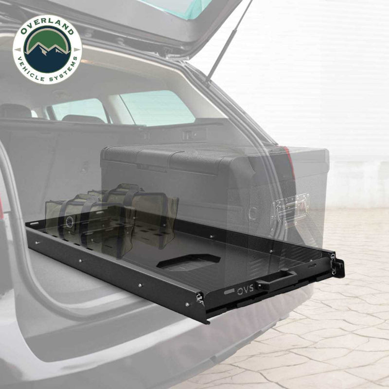 Image of an OVS black steel refrigerator slide extended from the back of a vehicle, with the cargo area of the vehicle visible, and a logo for "OVERLAND VEHICLE SYSTEMS" in the top left. This showcases the slide's practical application, making it easy to access items from the vehicle's rear