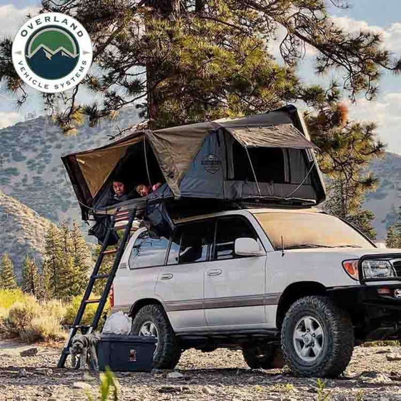 Image of OVS HD Bushveld II Hard Shell Roof Top Tent mounted on a white SUV, with a family inside the tent, ladder for access, and a dog in the foreground, set against a mountainous outdoor background