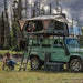Image of OVS HD Bushveld II Hard Shell Roof Top Tent mounted on a green SUV, with a person reading inside the tent, accessed by a ladder, and another person relaxing outside on a camping chair, set against a scenic outdoor backdrop