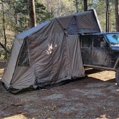 Image of the Overland Vehicle Systems (OVS) Hard Shell Bushveld tent, deployed with its annex room, attached to a gray Jeep. The setup is on a dirt surface surrounded by a forest