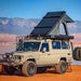 Image of Mamba 3 Clam Shell Roof Top Tent installed on a beige off-road vehicle, showing the tent fully deployed with a clear sky and mountain backdrop, angled front and side view