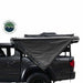 Image of an Overland Vehicle Systems Nomadic 180 Awning partially deployed on the side of a gray pickup truck, showing its sturdy dark gray fabric and aluminum frame. The awning provides shade over the rear side of the vehicle