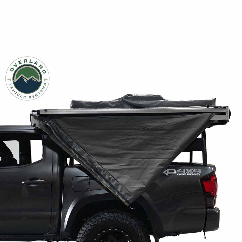 Image of an Overland Vehicle Systems Nomadic 180 Awning partially deployed on the side of a gray pickup truck, showing its sturdy dark gray fabric and aluminum frame. The awning provides shade over the rear side of the vehicle