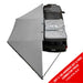 Image of OVS HD Nomadic 180 Degree Awning in dark grey with black travel cover, partially deployed on a vehicle, angled top view on white background