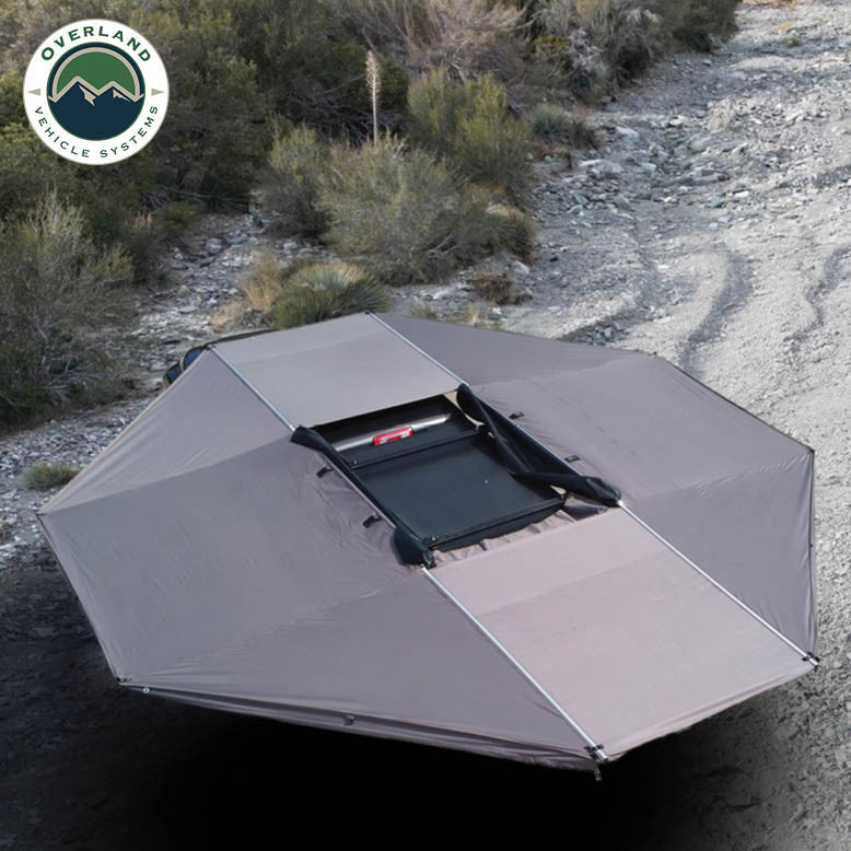 Image of OVS HD Nomadic 180 awning connector kit creating near-360-degree coverage around a vehicle, light gray fabric deployed in an outdoor off-road setting, angled top-down view.