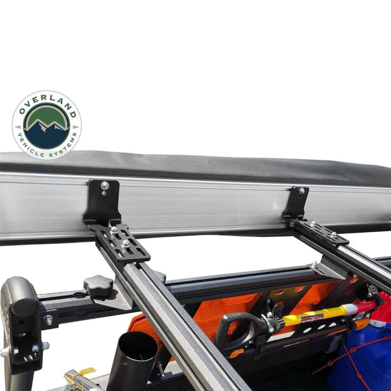 Image of the side view of an Overland Vehicle Systems 270-degree awning in its black travel cover, attached to a roof rack with two black mounting brackets. The aluminum awning casing is visible, as well as a yellow shovel and other gear mounted to the roof rack