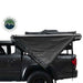 Image of an Overland Vehicle Systems 270-degree awning with walls, partially deployed from the side of a grey pickup truck. The grey awning fabric is angled down and backward, creating a triangular shape, and the black underside is visible