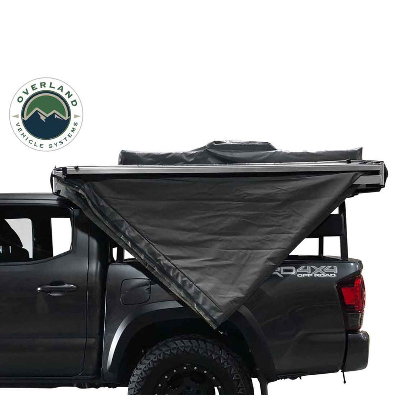 Image of an Overland Vehicle Systems 270-degree awning with walls, partially deployed from the side of a grey pickup truck. The grey awning fabric is angled down and backward, creating a triangular shape, and the black underside is visible