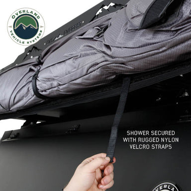 Image of an OVS HD Nomadic 2-person car-side shower and privacy room packed and secured on a roof rack using rugged nylon Velcro straps, shown in angled underside view on a white background.