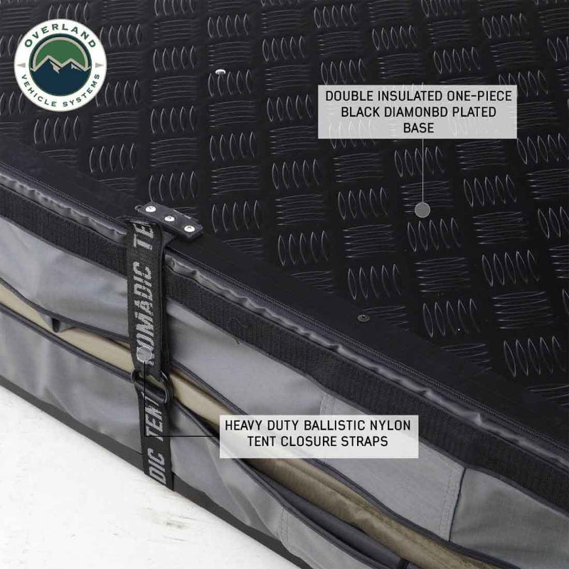 Image of a black diamond plate plated base of an Overland Vehicle Systems Nomadic Standard rooftop tent. The image highlights the double-insulated one-piece base and the heavy-duty ballistic nylon tent closure straps. The grey tent fabric with a zipper is visible, with a tent pole stored inside
