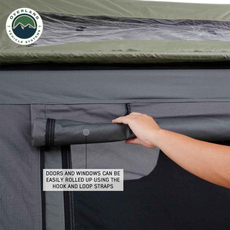Image shows a hand rolling up the grey window flap of an Overland Vehicle Systems Nomadic Standard rooftop tent, revealing the black mesh screen underneath. The text highlights that the doors and windows can be easily rolled up using hook and loop straps