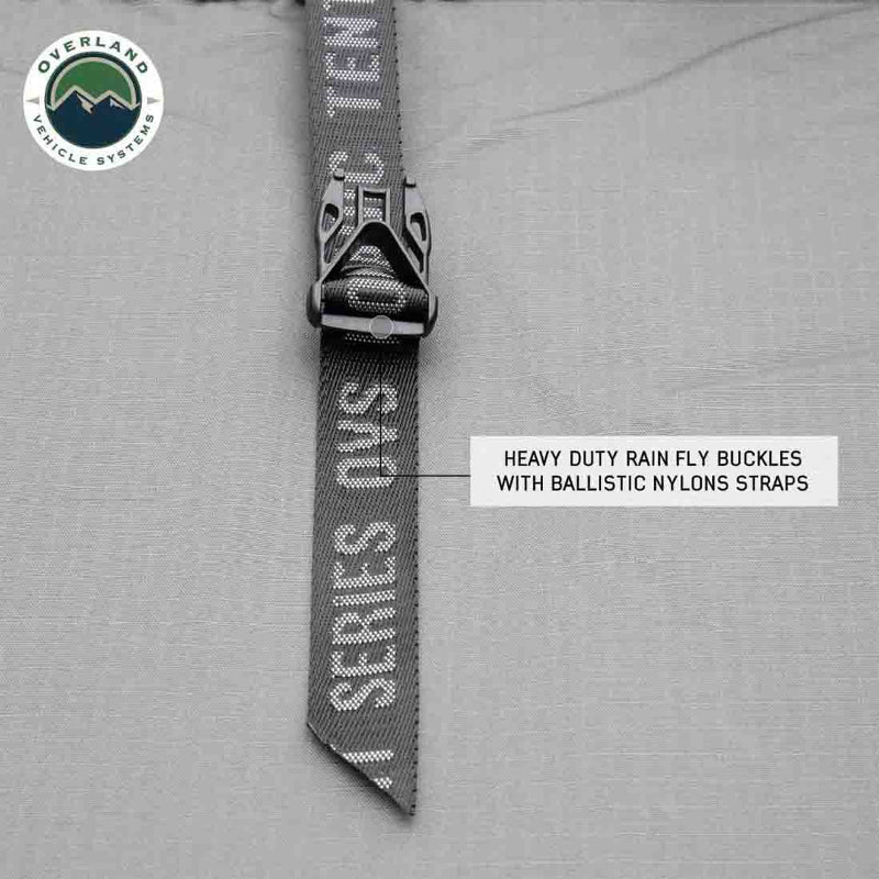 Image shows a close-up of a heavy-duty rain fly buckle with a ballistic nylon strap from an Overland Vehicle Systems product. The grey strap is emblazoned with "OVS NOMADIC SERIES TENT" and "OVERLAND VEHICLE SYSTEMS" logos