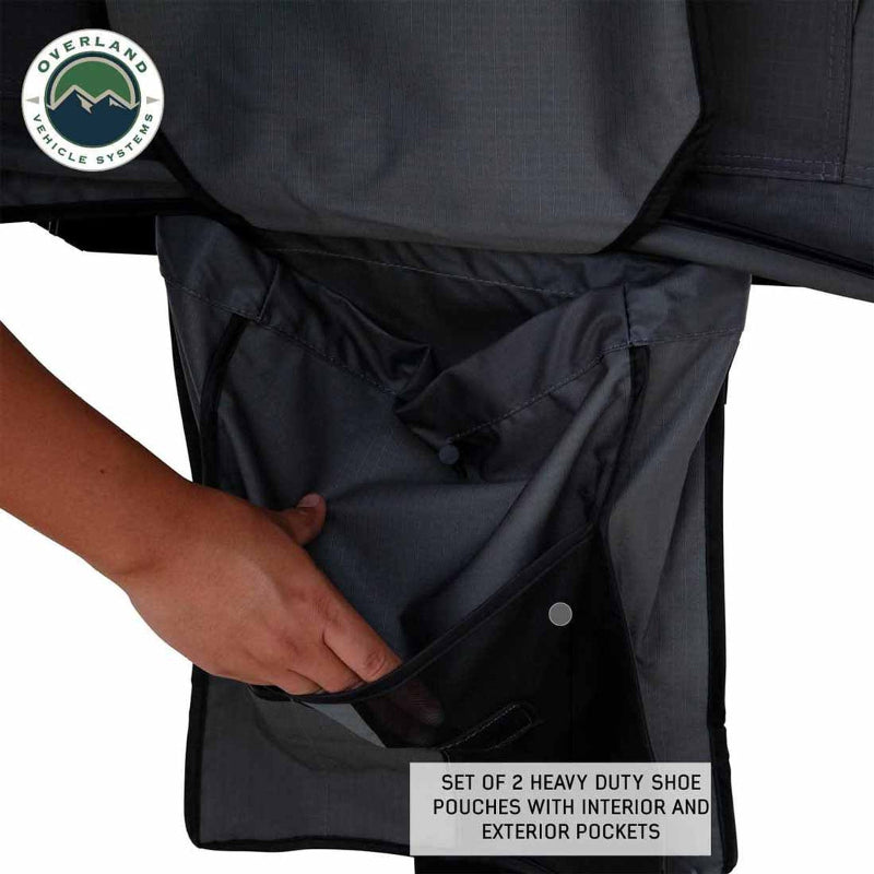 Image of a hand opening one of the two heavy-duty shoe pouches attached to a dark grey Nomadic 3 Extended roof top tent on a white background. The pouch has both interior and exterior pockets