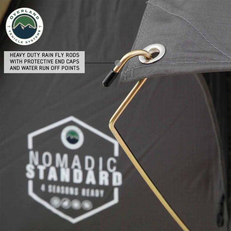 Image of a grey Overland Vehicle Systems Nomadic Standard awning, showing a gold support rod with a black capped end inserted into a silver grommet on the grey fabric. Text highlights "HEAVY DUTY RAIN FLY RODS WITH PROTECTIVE END CAPS AND WATER RUN OFF POINTS" and the awning features a white logo with "NOMADIC STANDARD 4 SEASONS READY