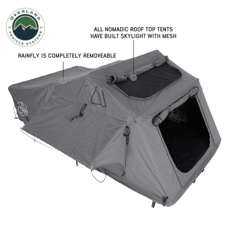 Image displaying a dark grey Nomadic Roof Top Tent, fully deployed with a rainfly that has a built-in skylight and mesh windows, shown at an angle with the rainfly indicated as completely removable. The "Overland Vehicle Systems" logo is in the top left corner, all on a white background