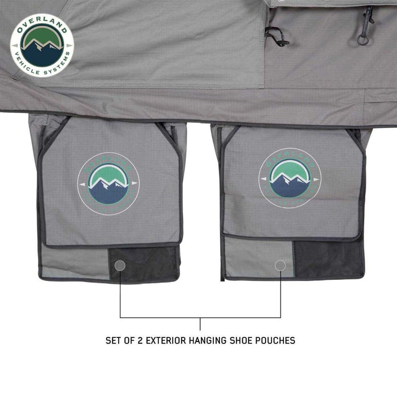 Image showcasing a pair of grey exterior hanging shoe pouches with the "Overland Vehicle Systems" mountain logo, positioned underneath a darker grey tent fabric. The "Overland Vehicle Systems" logo is in the top left corner