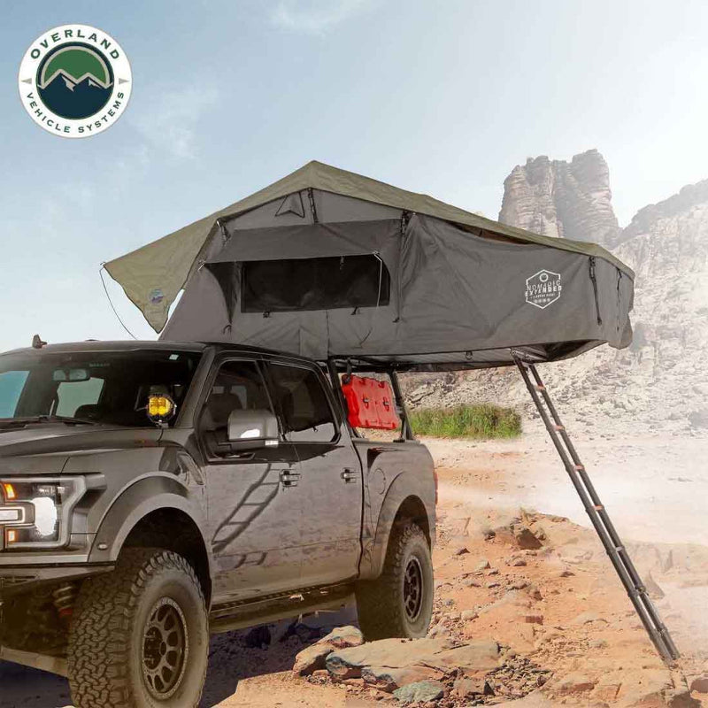 Image of a dark grey hardshell rooftop tent, partially deployed with an awning extended, mounted on a grey pickup truck in a rugged, mountainous desert landscape. A ladder extends from the tent to the ground, with the "Overland Vehicle Systems" logo visible in the top left corner