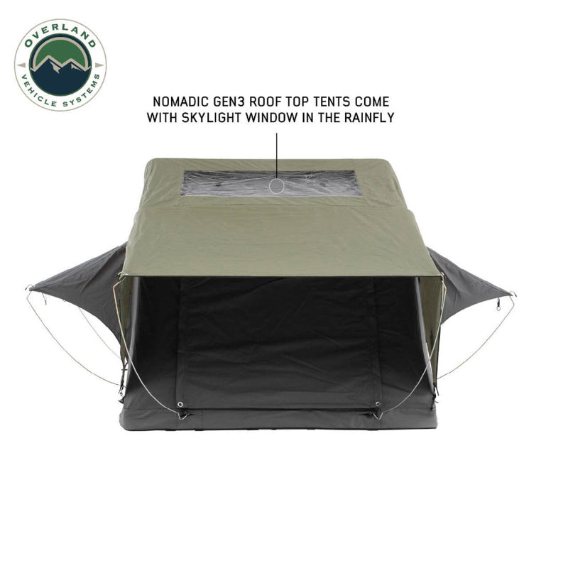 Image of an olive green Nomadic Gen3 Roof Top Tent, fully deployed with a clear skylight window in the rainfly, shown from the front with guy lines extended. The "Overland Vehicle Systems" logo is in the top left corner, all on a white background