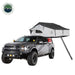 Image of a grey Ford pickup truck with a white and dark grey Nomadic Extended rooftop tent fully deployed on its roof, including an attached ladder extending to the ground, set against a white background